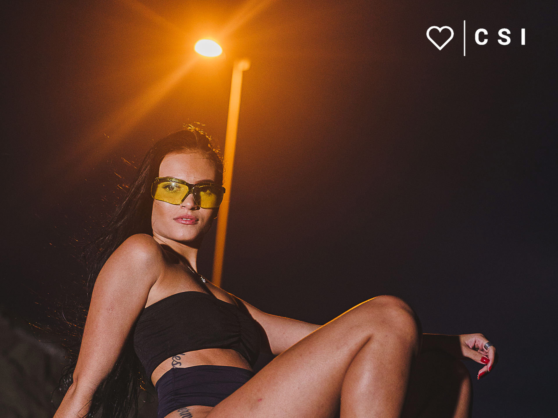 modelo fotografica com look preto sentada sobre uma pedra com oculos tatico marelo a noite com poste amarelo ao fundo