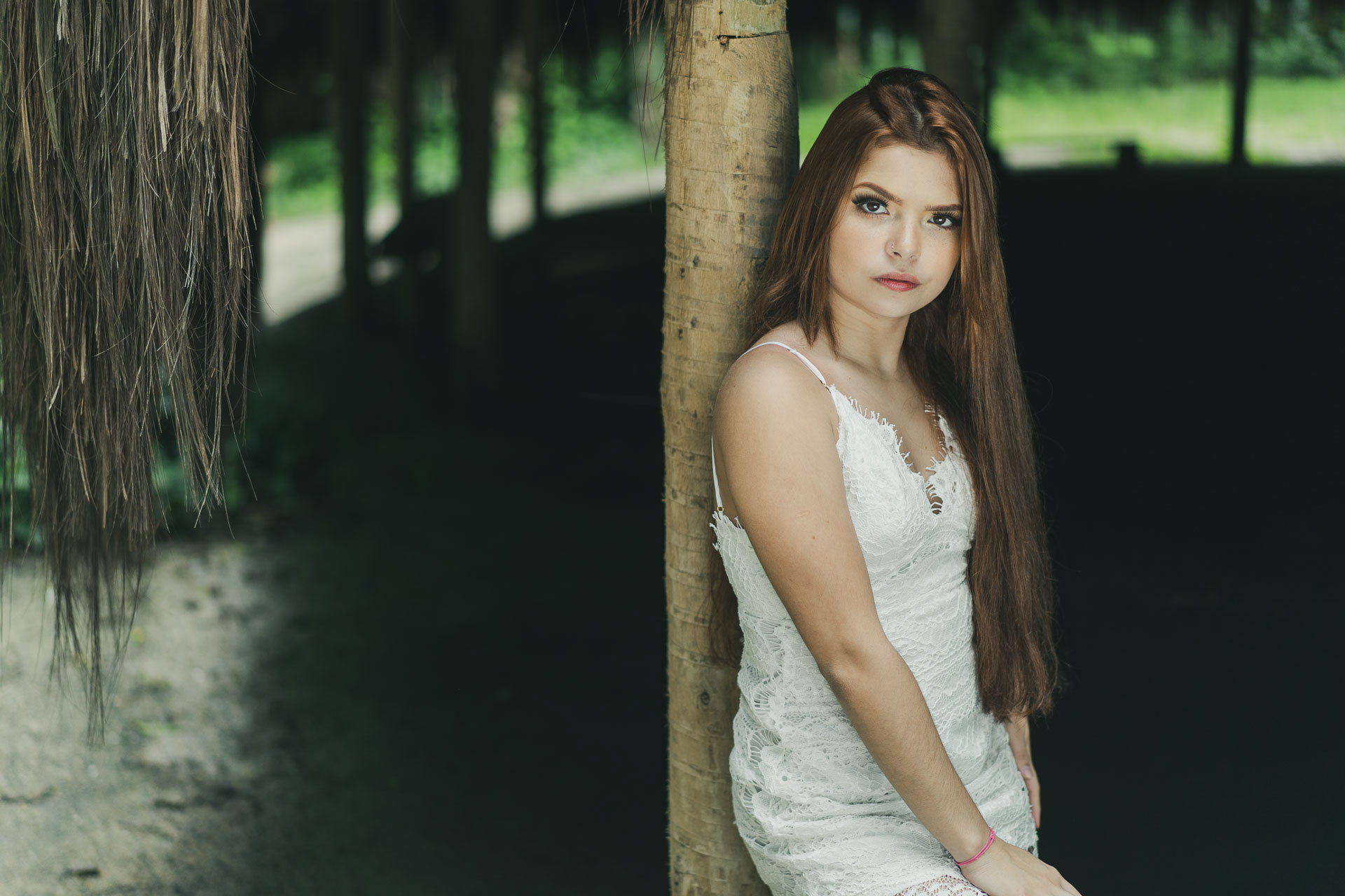 modelo ruiva de vestido de renda branca apoiada no tronco de madeira da oca do parque laje com o fundo escuro aparencia séria e cabelo longo